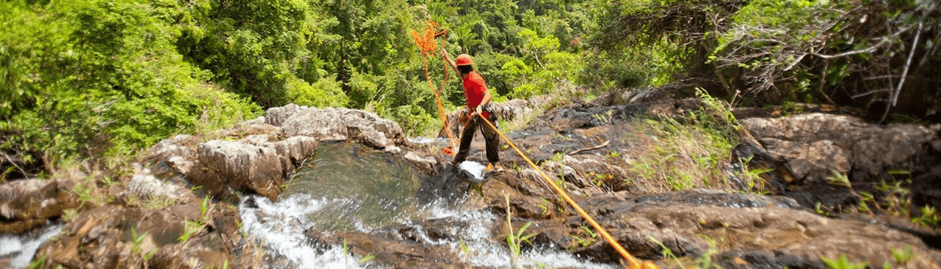 A man in an orange helmet and tee shirt rappels down a waterfall in a jungle setting.