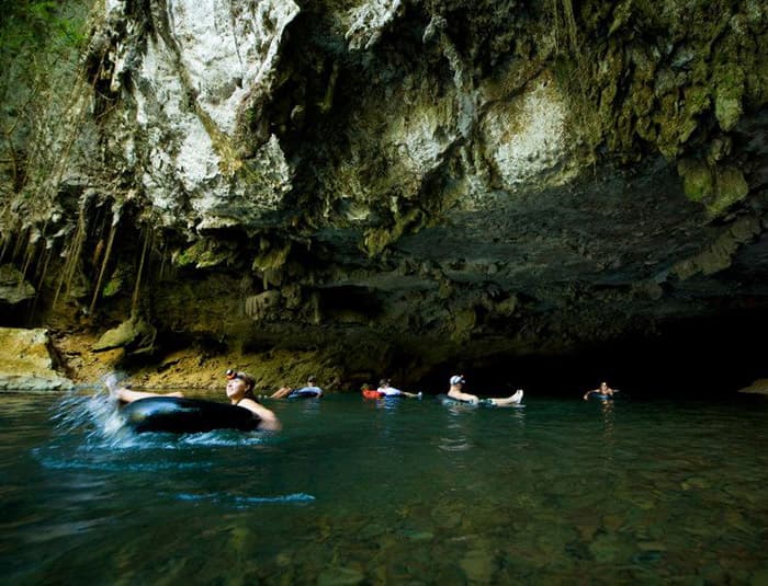 A group of people are tubing down a dark underground river in a cave. The water is calm and clear, and stalactites hang from the ceiling.
