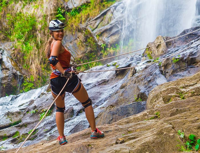 A young woman in an orange tank top smiles over her shoulder as she gets ready to rappel down rocks next to a waterfall.