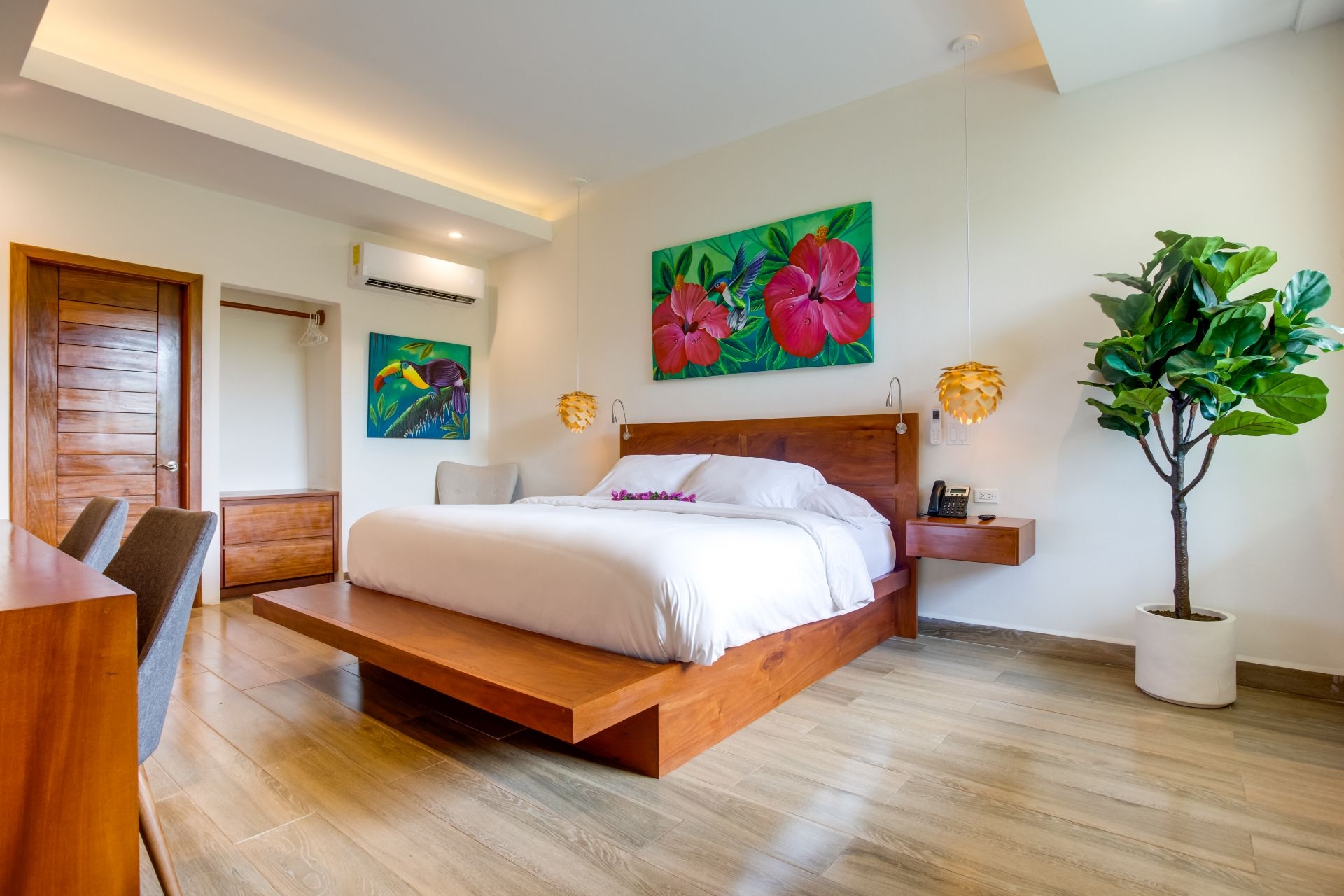 The image shows a hotel room with a modern design. The room features a large bed with white linens, a wooden accent wall with a built-in TV, a wooden desk with two chairs, a colorful painting on the wall, and a large window with natural light.