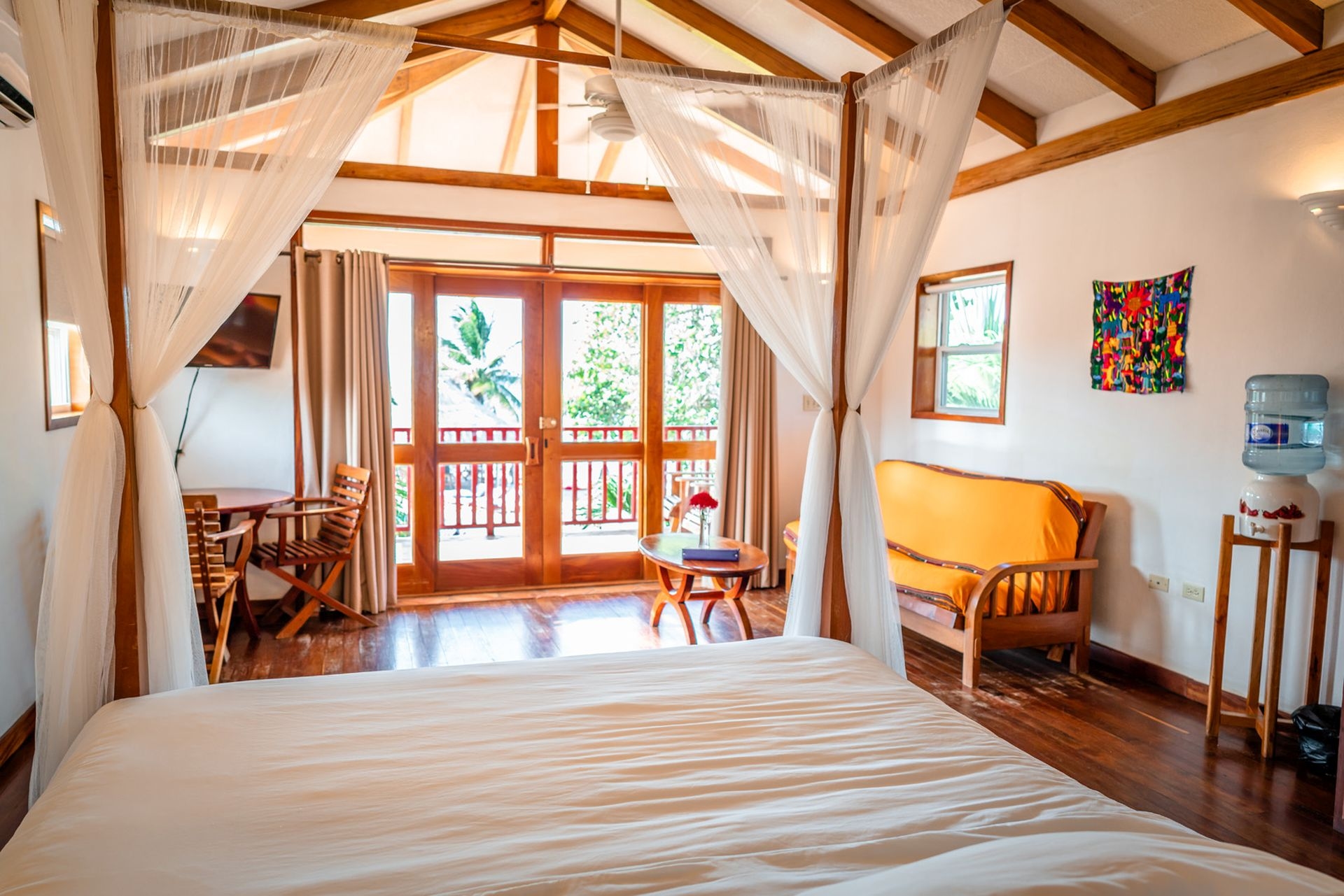 A spacious bedroom with a four-poster bed covered in white sheets. The bed has sheer white curtains hanging from the canopy. A ceiling fan hangs from the center of the room, and there is an air conditioning unit in the background. Nightstands with lamps flank the bed, and a wooden bench sits at the foot. A small sofa sits in the corner of the room, and a coffee table is placed between the sofa and the bed. Sliding glass doors lead to a balcony overlooking a view of the ocean. A colorful tapestry hangs on the wall behind the bed.