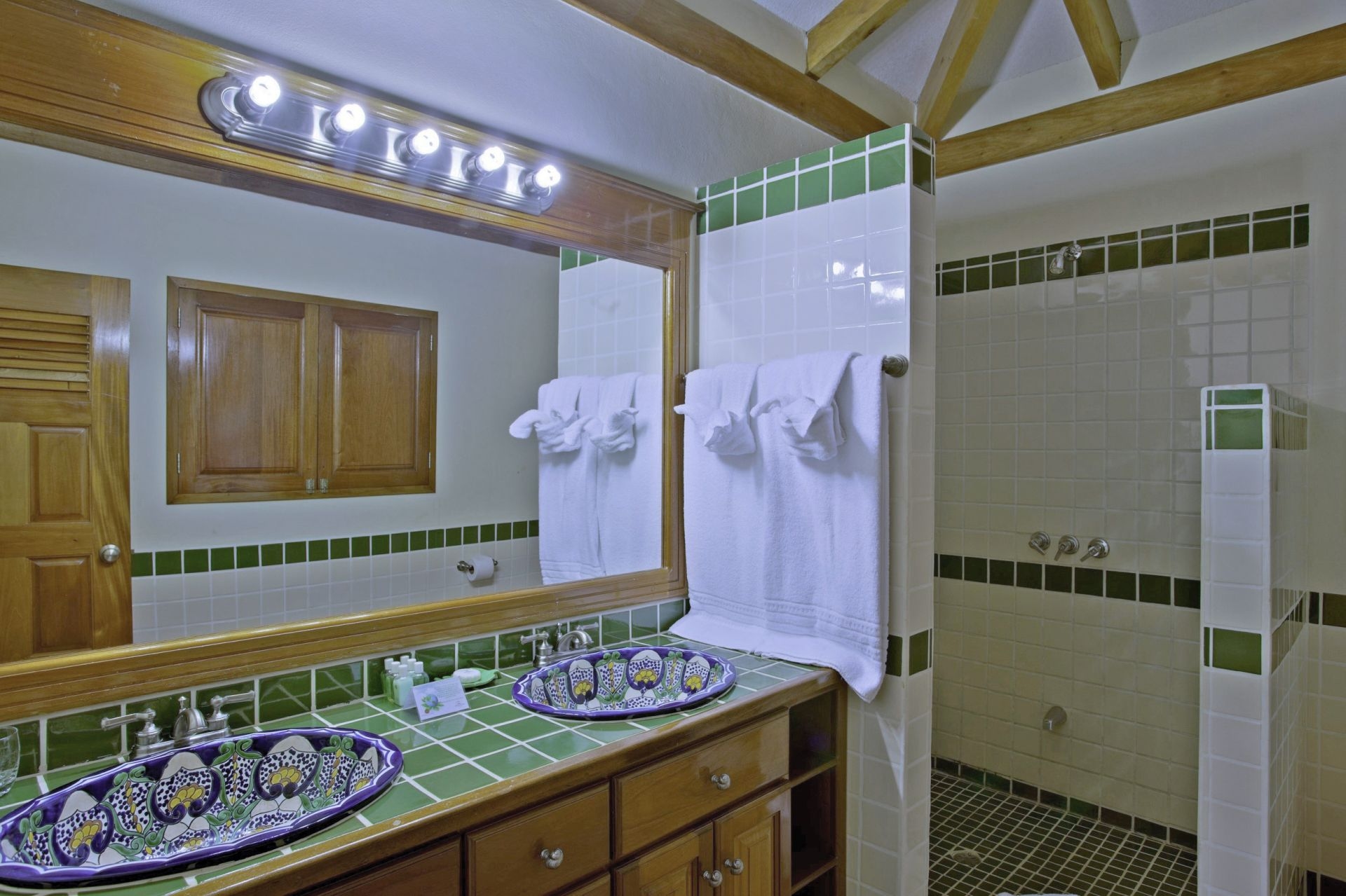 A spacious bathroom with dual sinks, a large mirror, and a walk-in shower. The walls are tiled with green and white tiles, and the floors are made of stone. There is a wooden vanity with drawers underneath the sinks, and a shelving unit with towels and other toiletries. The bathroom has a bright and airy feel with plenty of natural light. The sinks have colorful, patterned bowls.