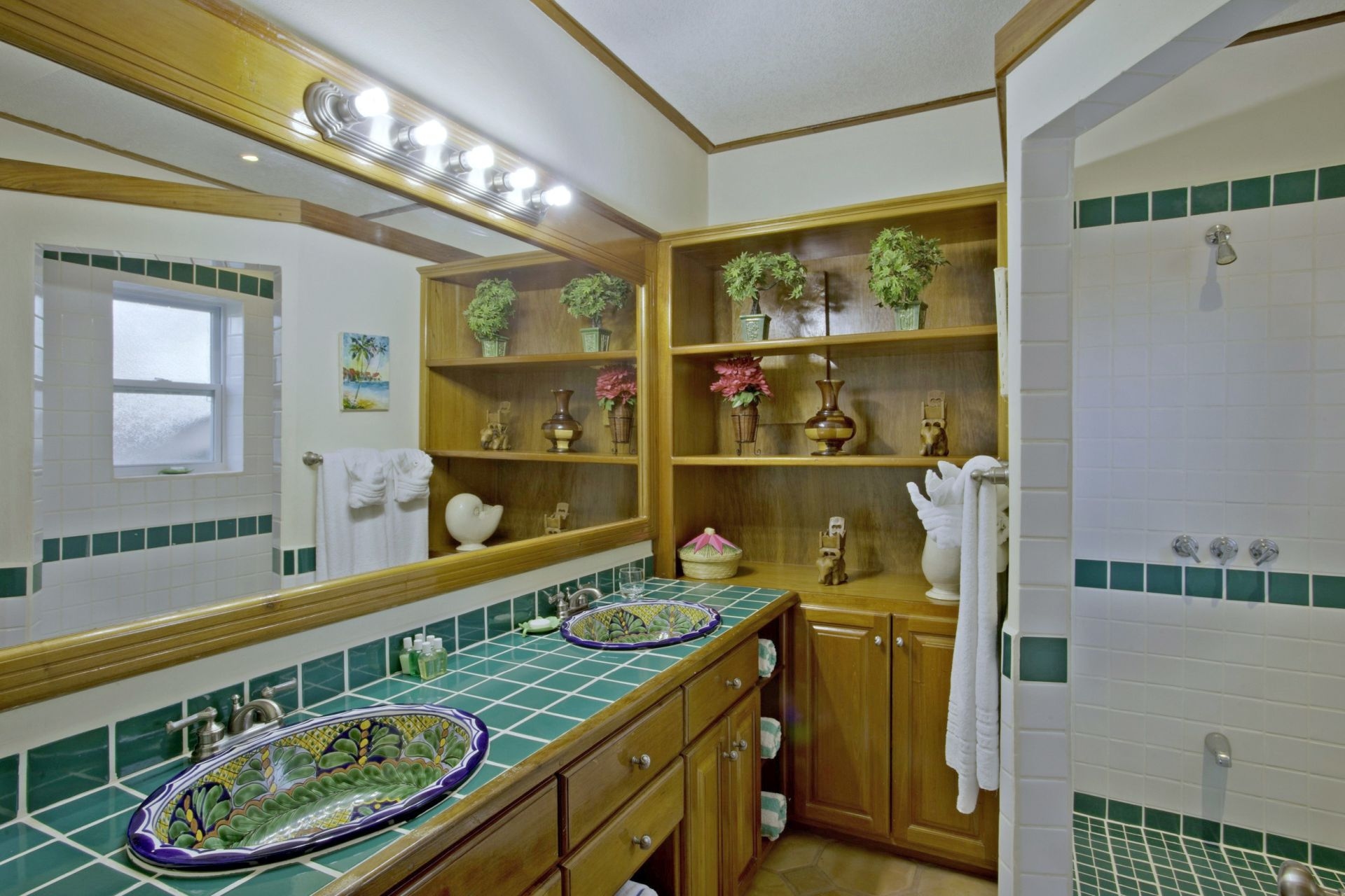 A spacious bathroom with dual sinks, a large mirror, and a walk-in shower. The walls are tiled with green and white tiles, and the floors are made of stone. There is a wooden vanity with drawers underneath the sinks, and a shelving unit with towels and other toiletries. The bathroom has a bright and airy feel with plenty of natural light. The sinks have colorful, patterned bowls.