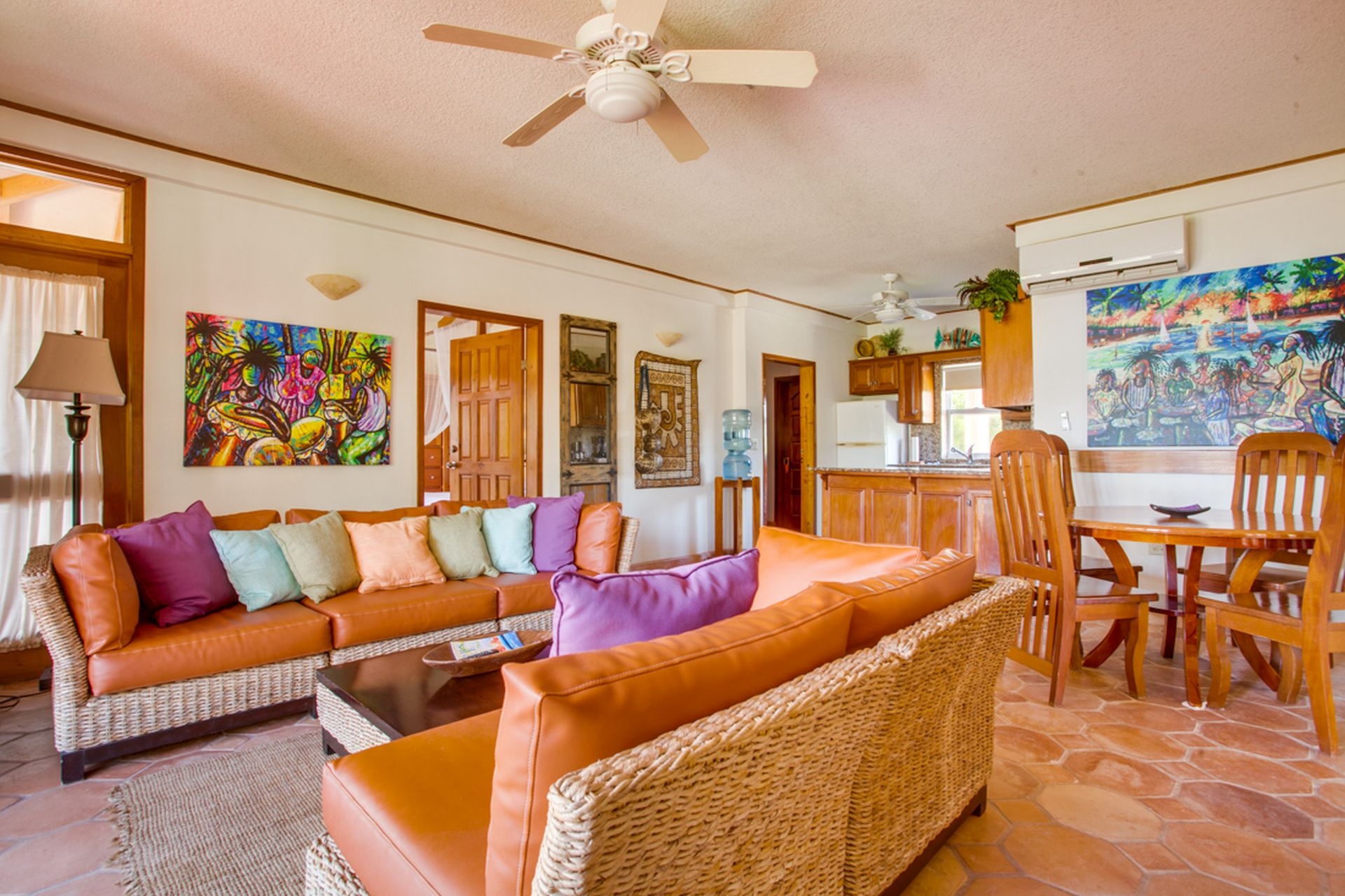 A spacious, brightly lit living room with a comfortable leather couch, colorful throw pillows, and wicker armchairs. A large ceiling fan hangs from the center of the room, and there are two air conditioning units in the background. A dining table with chairs is visible in the corner, and a kitchen area is partially shown on the right side of the image. Two pieces of colorful artwork hang on the walls.