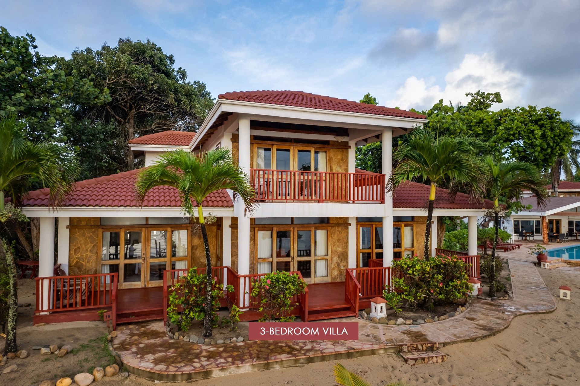 The image shows a 3-bedroom villa at Belizean Dreams Resort in Hopkins, Belize. The villa is a two-story building with a red tile roof and a large balcony on the second floor. The villa is located on the beach and surrounded by palm trees and lush greenery.