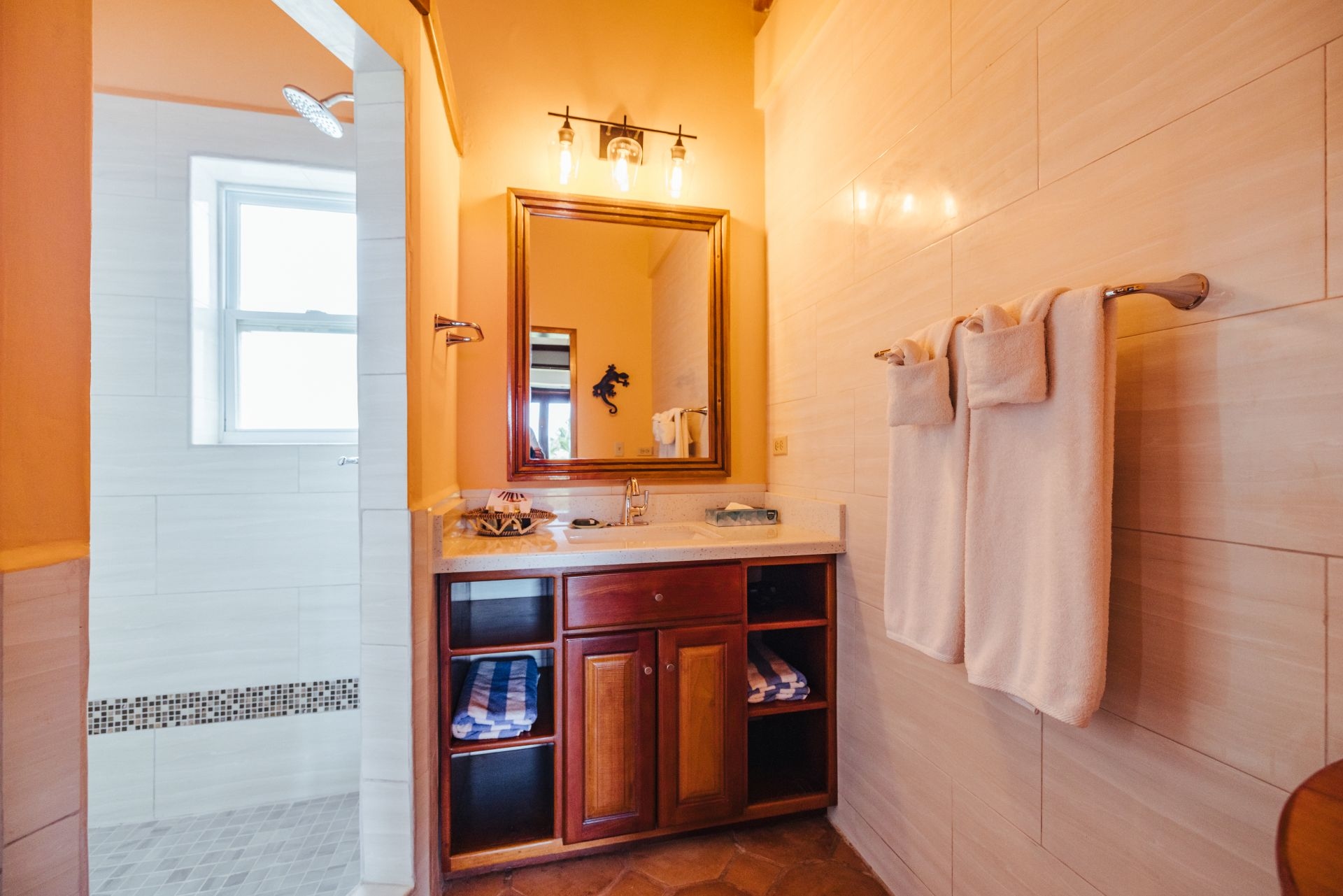 A spacious bathroom with a single sink, a large mirror, and a walk-in shower. The walls are tiled, and the floors are made of stone. There is a wooden vanity with drawers underneath the sink, and a shelving unit with towels and other toiletries. The bathroom has a bright and airy feel with plenty of natural light.