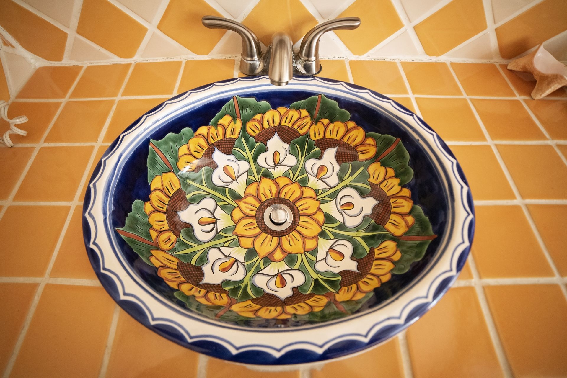 A circular ceramic sink with a colorful Mexican Talavera design featuring sunflowers, lilies, and green leaves. The sink is set against a backdrop of yellow and orange square tiles.