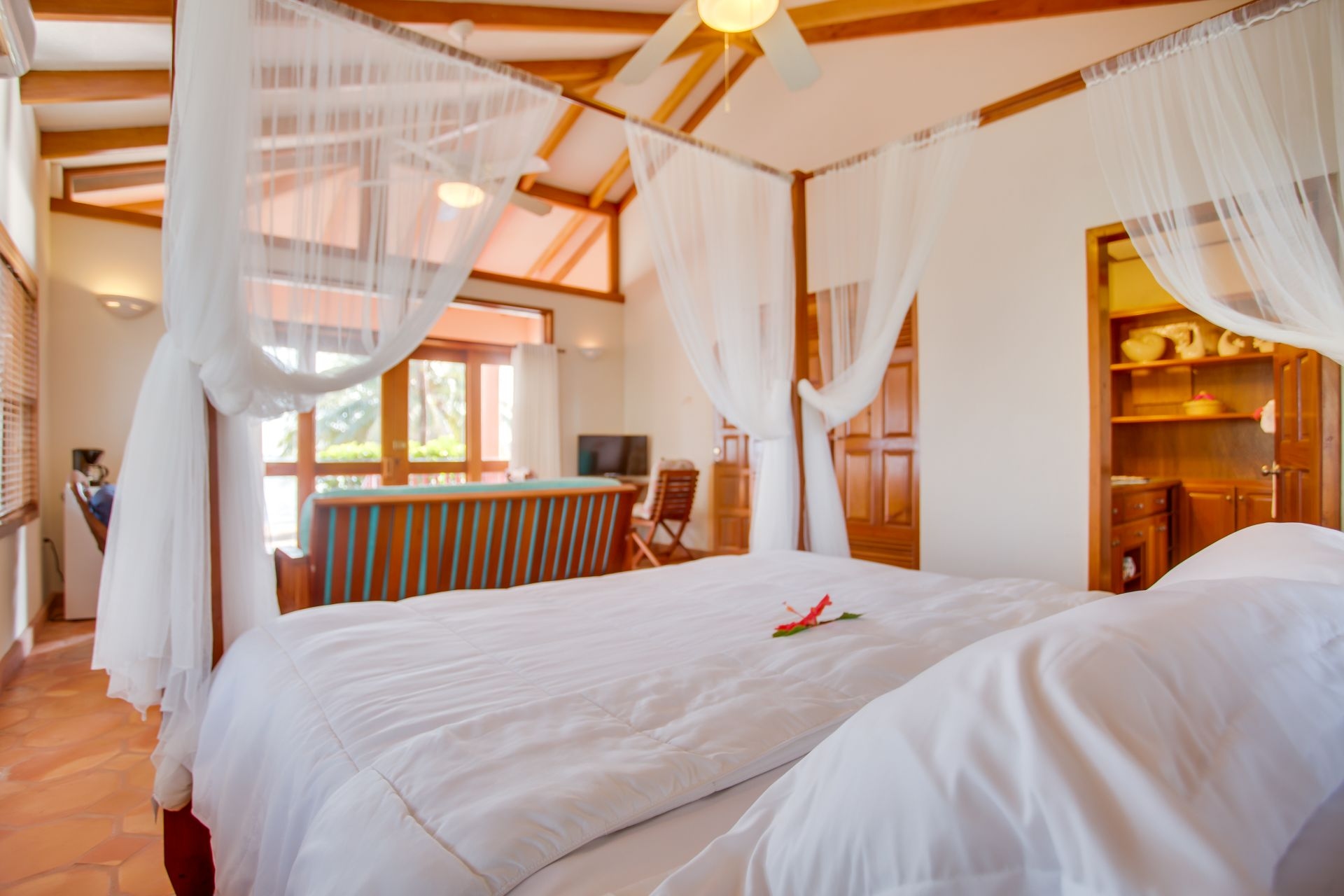 A spacious bedroom with a four-poster bed covered in white sheets. The bed has sheer white curtains hanging from the canopy. A ceiling fan hangs from the center of the room, and there is an air conditioning unit in the background. Nightstands with lamps flank the bed, and a wooden bench sits at the foot. A small sofa sits in the corner of the room, and a coffee table is placed between the sofa and the bed. Sliding glass doors lead to a balcony overlooking a living area with a couch and chairs. A colorful painting hangs on the wall behind the bed.