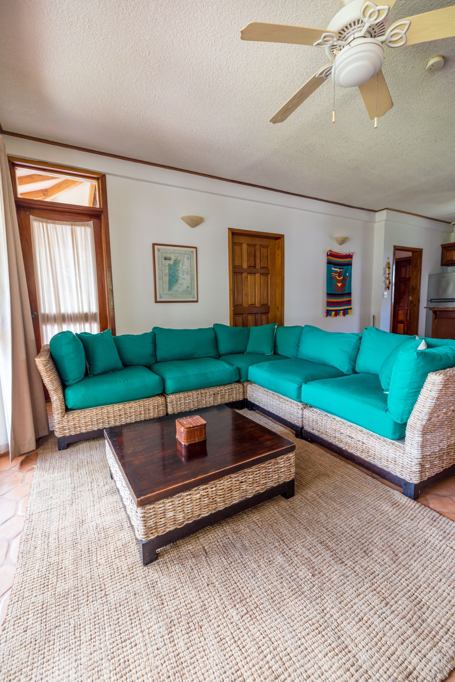 A spacious, brightly lit living room with a large, teal-colored sectional sofa, a wicker coffee table, and a woven rug. A ceiling fan hangs from the center of the room, and there are air conditioning units in the background. A door leads to a balcony or patio, and a window provides natural light.