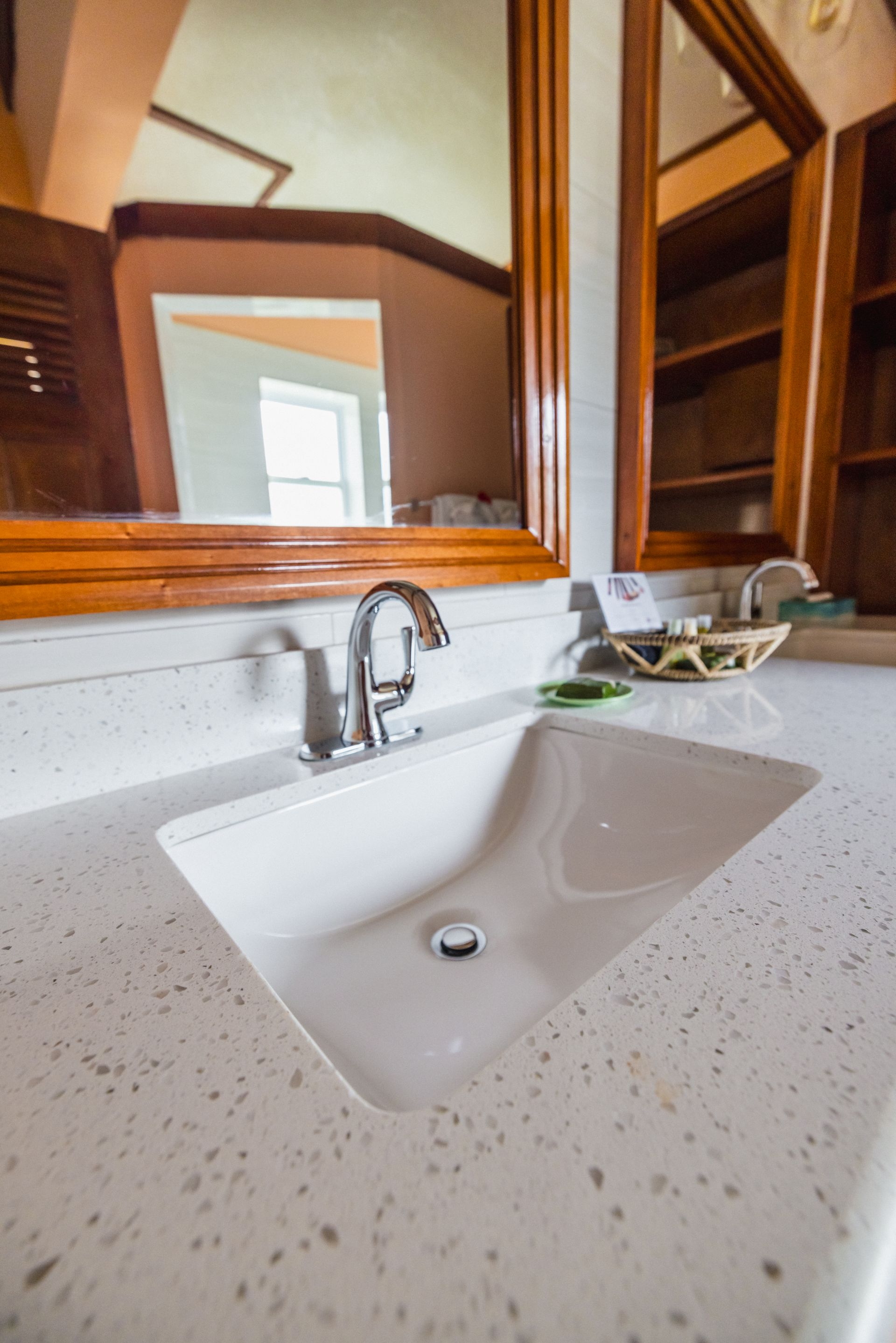 A close-up of a white rectangular sink with a chrome faucet. The sink is set on a white speckled countertop, and there is a wooden framed mirror above it. A small basket with toiletries sits next to the sink.