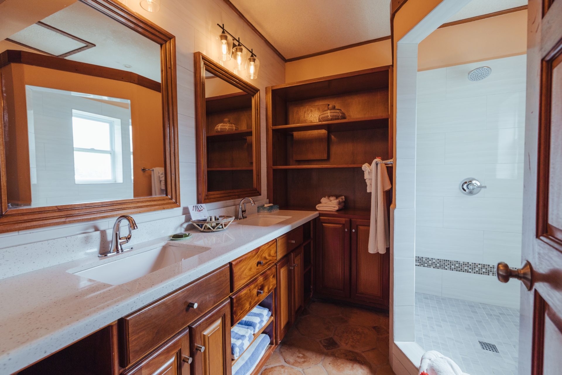 A spacious bathroom with dual sinks, a large mirror, and a walk-in shower. The walls are tiled, and the floors are made of stone. There is a wooden vanity with drawers underneath the sinks, and a shelving unit with towels and other toiletries. The bathroom has a bright and airy feel with plenty of natural light.