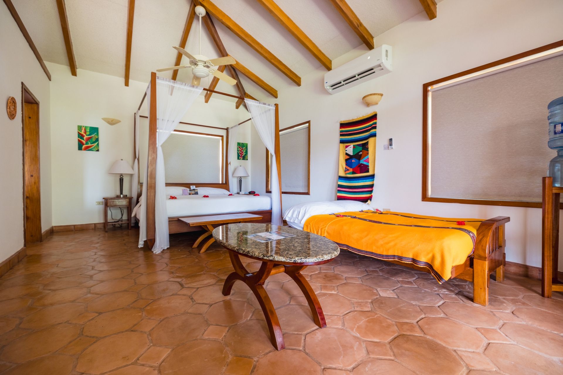 A spacious bedroom with a four-poster bed covered in white sheets. The bed has sheer white curtains hanging from the canopy. A colorful tapestry hangs on the wall behind the bed, and a wooden bench sits at the foot. The room has a tiled floor and a wooden nightstand with a lamp.