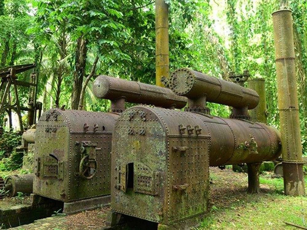 A group of rusty, overgrown sugar mill machines in a dense jungle setting. The machines appear to be boilers or steam engines, with pipes and valves visible.