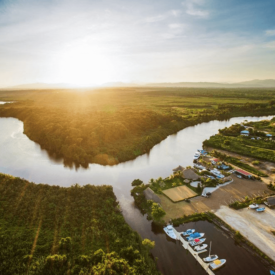 An aerial view of a river winding through a lush, tropical landscape. A marina with boats is visible on the shore, and the sun is setting over the horizon, casting a golden glow on the scene.