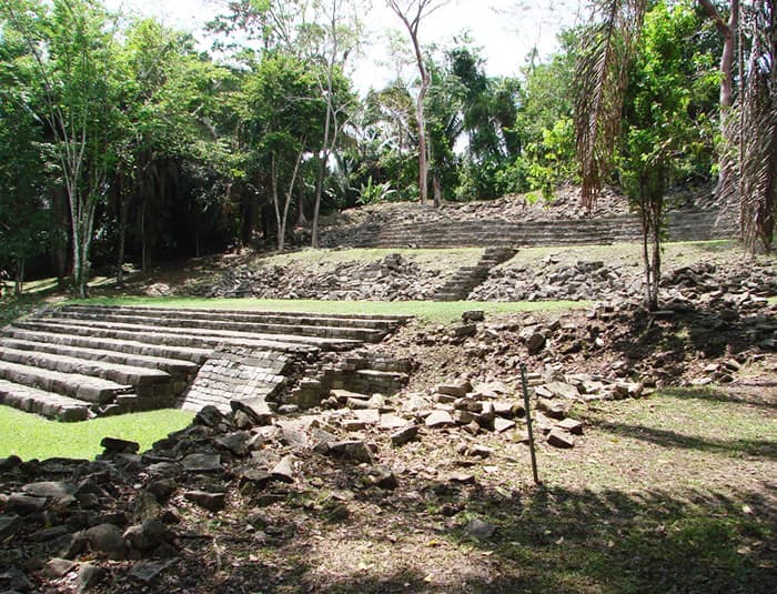 A photo of the Lubaantun Mayan ruins in Belize. The image shows a large, stepped platform made of stone, surrounded by lush green vegetation and trees.