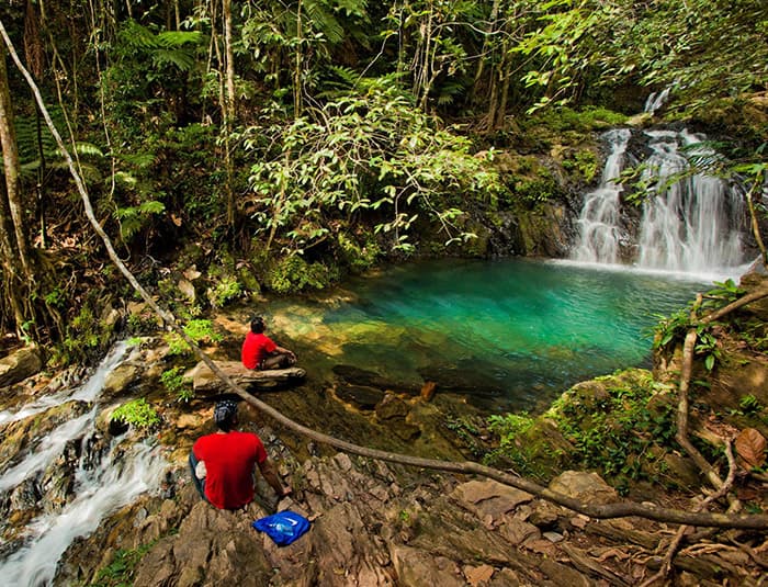 Two people in red shirts are sitting on rocks overlooking a beautiful turquoise pool surrounded by lush green rainforest. A small waterfall cascades into the pool.