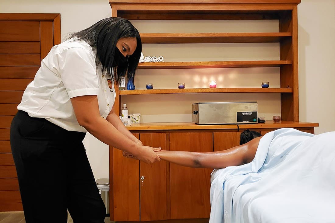 A masked massage therapist gives a leg massage to a spa guest on a white-draped table.