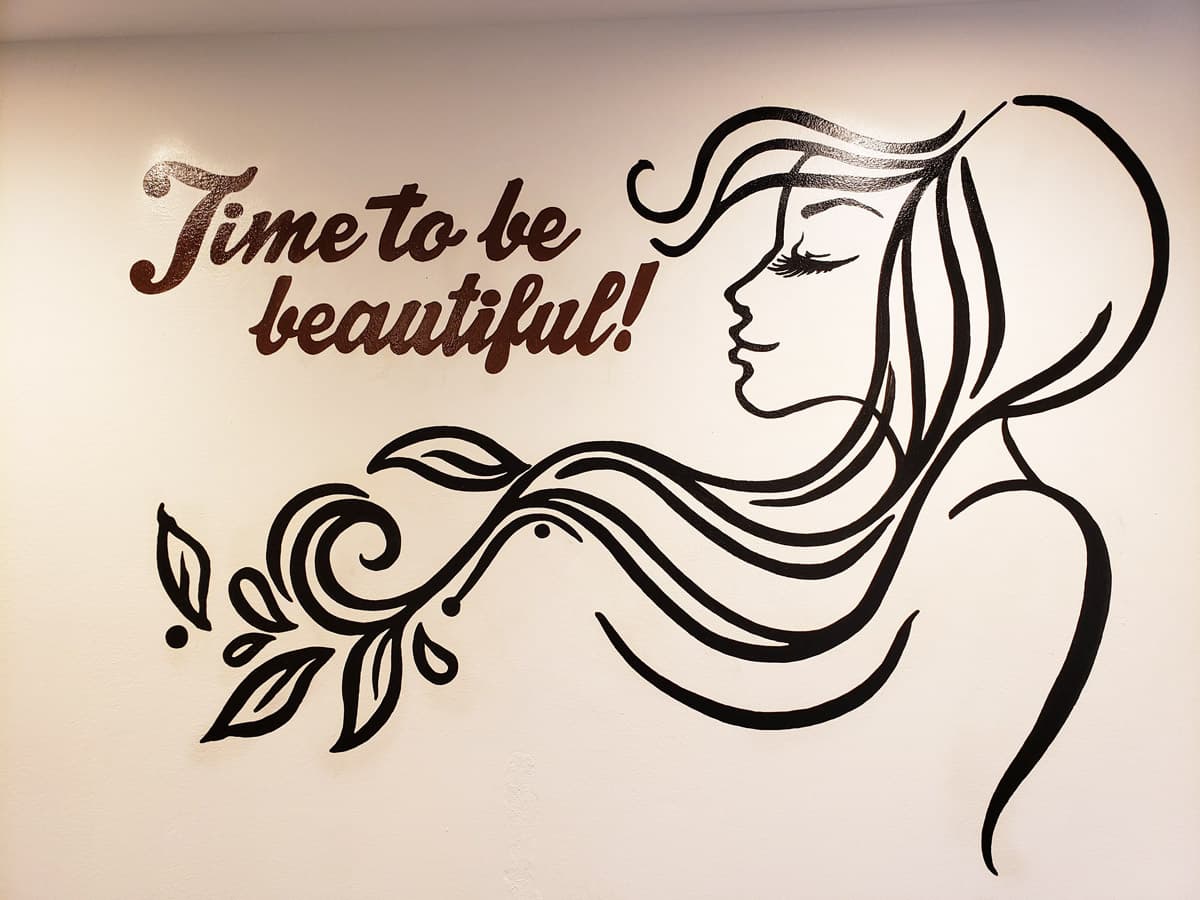 A wall decal with the text "Time to be beautiful!" and a side profile of a woman with flowing hair. The woman's hair is stylized in a wavy pattern and appears to be blowing in the wind. There is a small floral design below the woman's face. The decal is black and white, with the text in a cursive font.