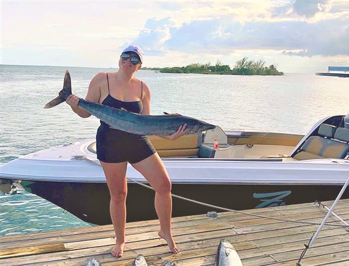 A woman proudly holds a long gray fish on a dock in front of a nice boat.