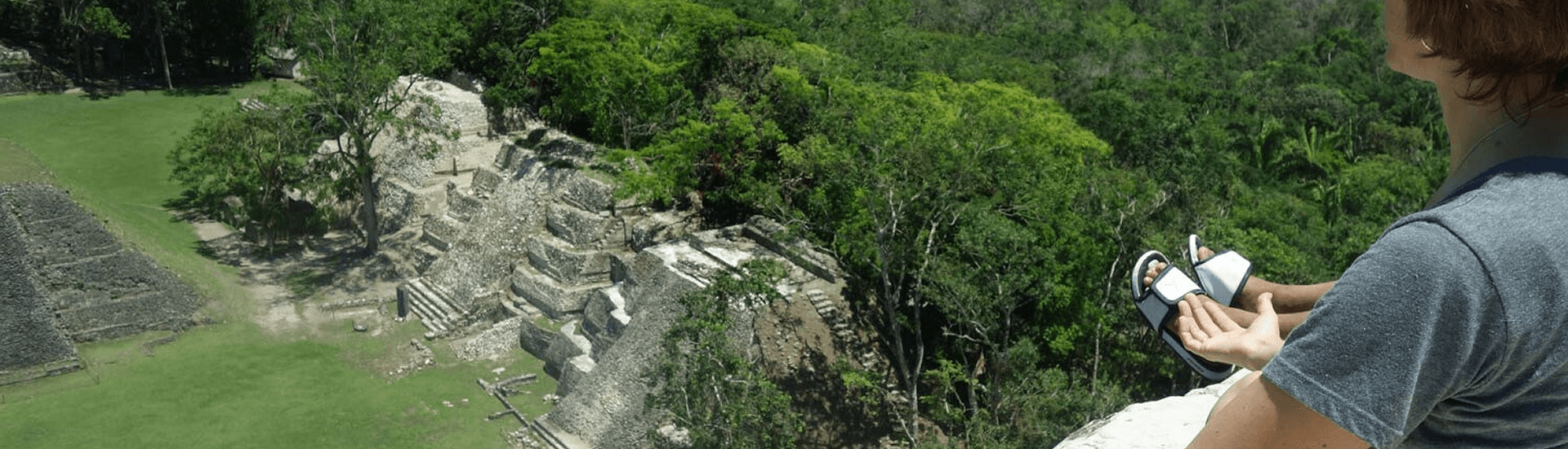 A woman sits on a high level overlooking a mayan ruin in the jungle.