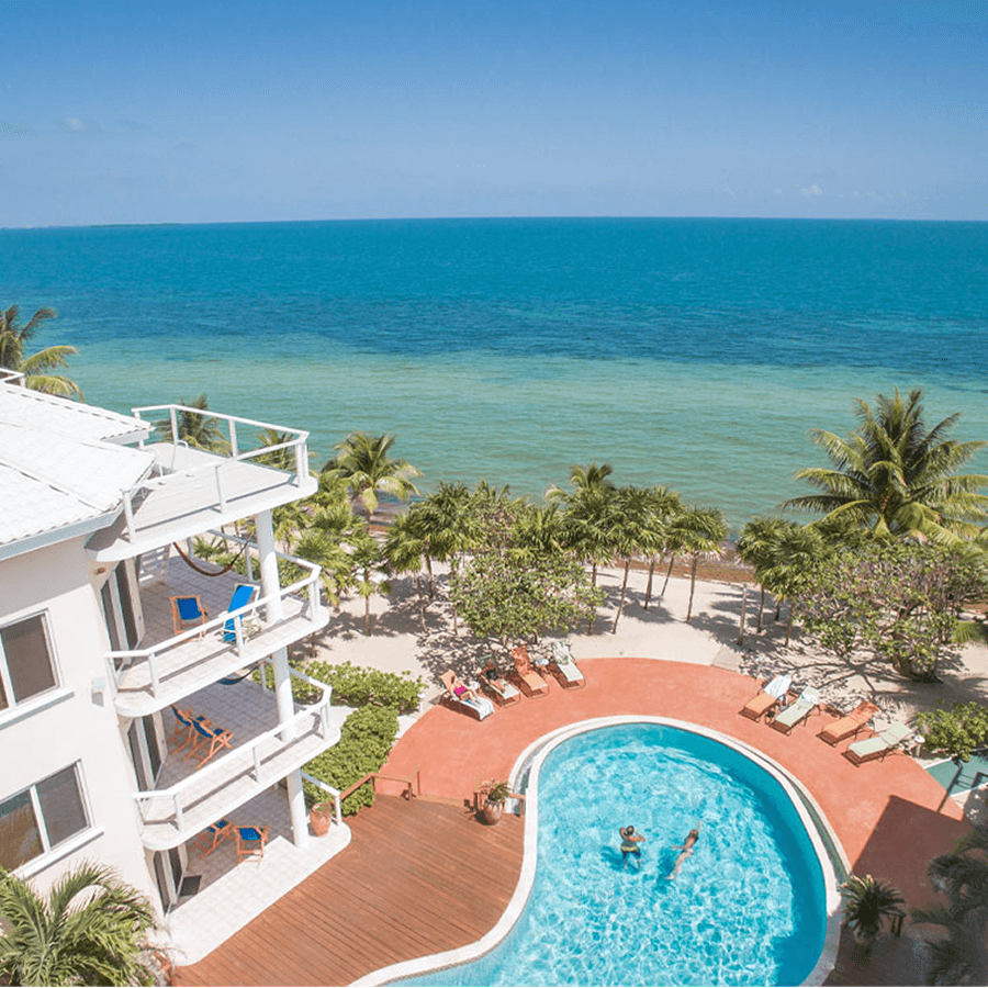 An aerial view of a beachfront resort with white buildings, a large swimming pool, and palm trees. The ocean is visible in the background.