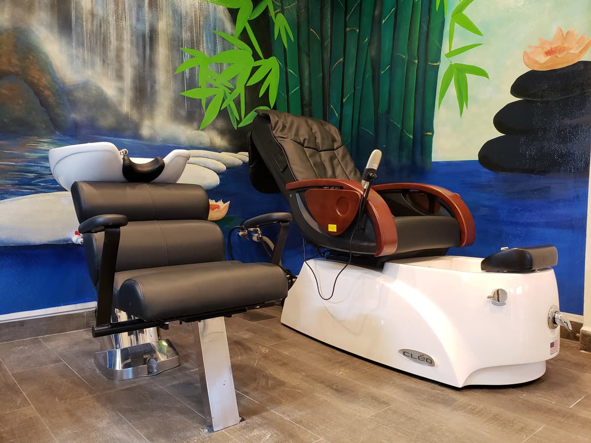 A spa setting with a large mural depicting a waterfall and bamboo forest. In the foreground, a black shampoo chair sits next to a white pedicure station with a massage chair. Lotus flowers float on the water at the base of the waterfall. The spa has a modern aesthetic with sleek black and white furniture and large mirrors.