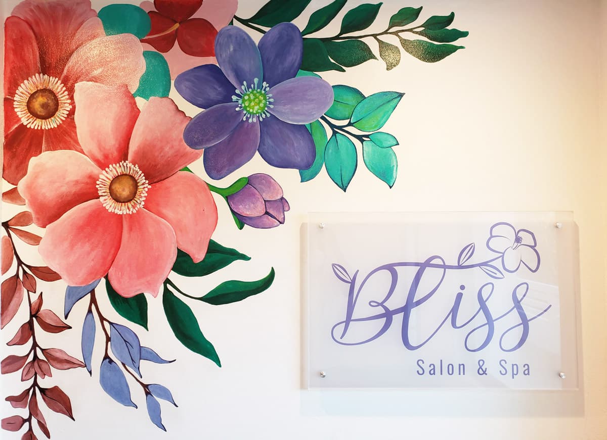 A vibrant mural of colorful flowers adorns a wall, with a clear acrylic sign for "Bliss Salon & Spa" positioned prominently beside it. The mural features a variety of blooms in shades of pink, purple, and blue, with green leaves and branches adding depth. The salon's name is written in a flowing script on the sign, with a smaller "Salon & Spa" tagline beneath it.