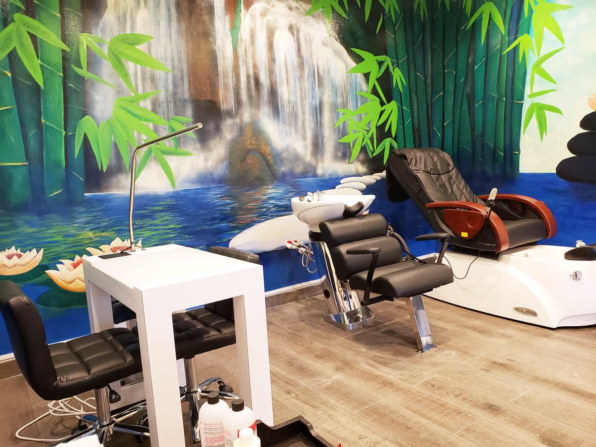 A serene spa setting with a large mural depicting a waterfall and bamboo forest. The mural is painted on a white wall behind a shampoo station and a massage chair. Lotus flowers float on the water at the base of the waterfall. The spa has a modern aesthetic with sleek black and white furniture and large mirrors.