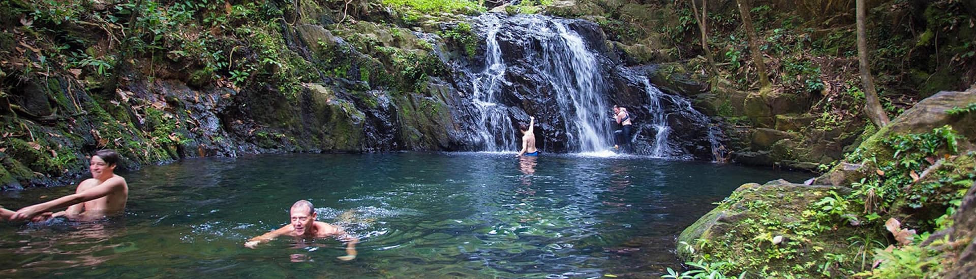 Multiple men swim through the water as a man and a woman climb up to dive off of a waterfall in the background.