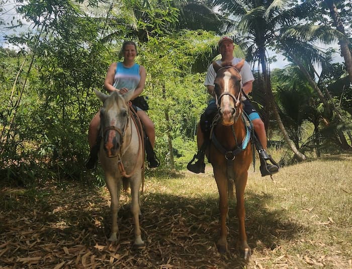 A couple ride white and brown horses through the jungle.