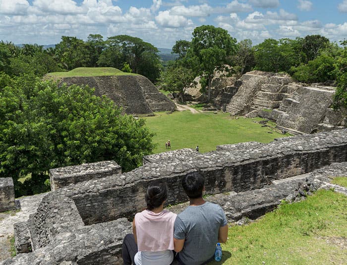 A young couple sits overlooking a mayan ruin, surrounded by stone walls and jungle foliage.