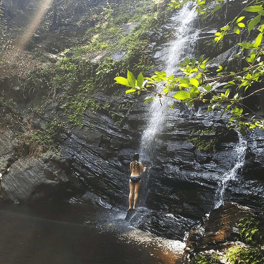 A woman in a bikini is standing under a waterfall in a lush, green forest. The water cascades down a rocky cliff and into a pool below.