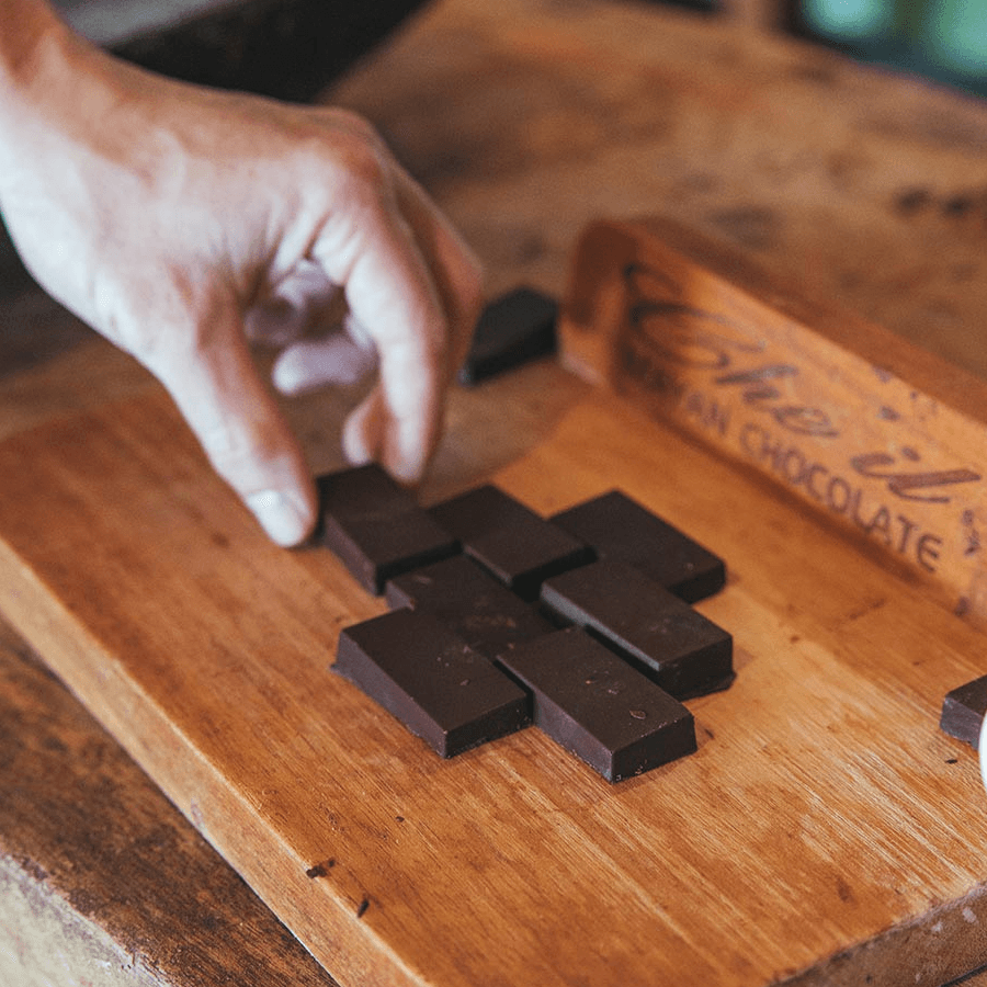 A person's hand arranging small squares of dark chocolate on a wooden cutting board. The chocolate has a glossy finish and is surrounded by a wooden box labeled "Artisan Chocolate."