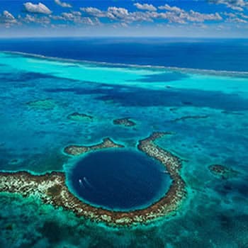 An aerial view of the Great Blue Hole in Belize. It's a circular, dark blue hole surrounded by a vibrant coral reef and turquoise ocean. The sky is clear with white clouds.