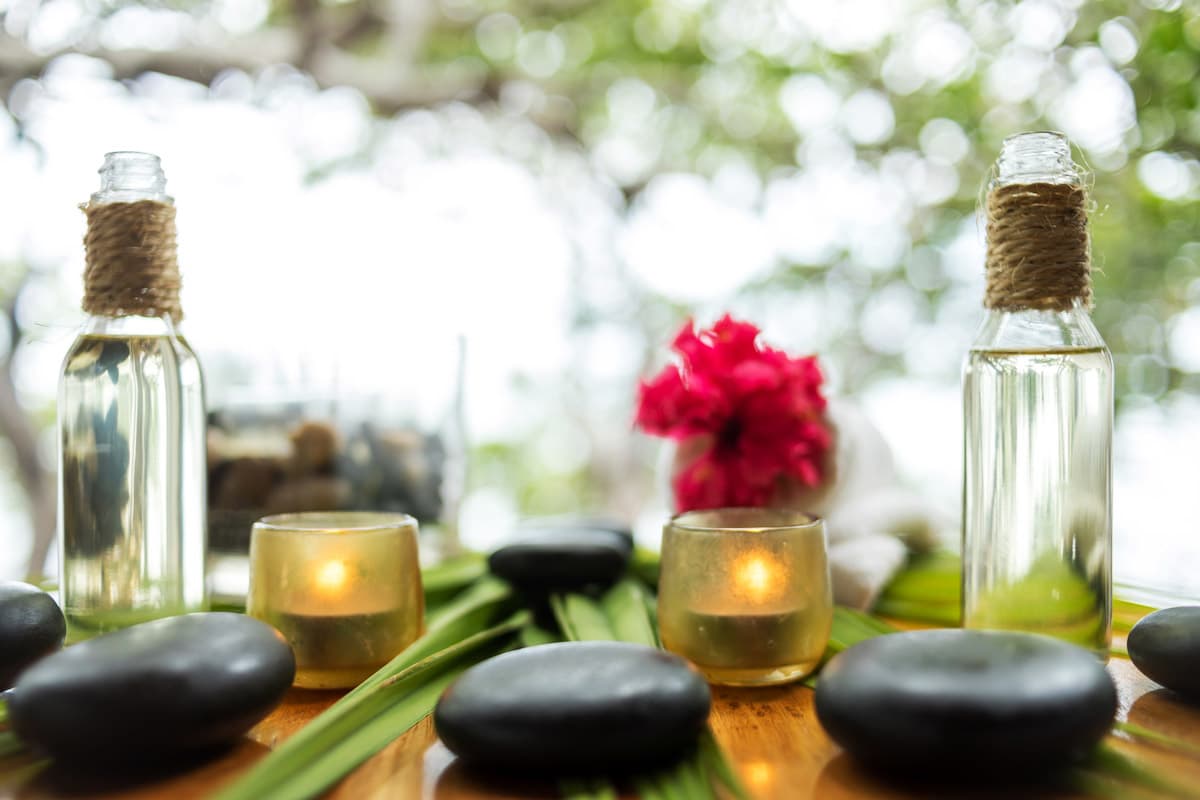 A spa-like setting with two bottles of essential oil, a small potted plant, two lit candles, and several smooth, black stones arranged on a wooden surface. The bottles are made of clear glass and are tied with twine. The potted plant is a small, red flower. The candles are placed in small, glass containers. The stones are arranged in a group, with some placed on top of green leaves. The background is blurred, but it appears to be a natural setting, possibly outdoors.