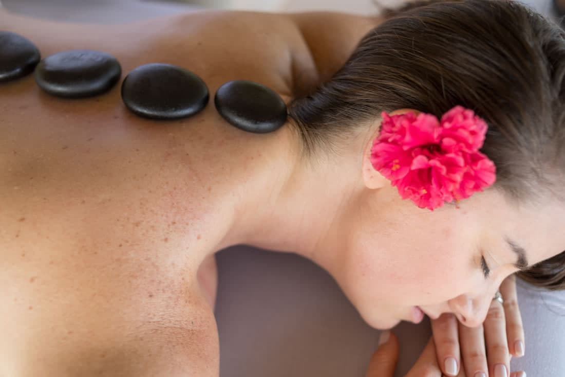 A brunette woman with a pink flower in her hair receives a hot stone massage spa service.