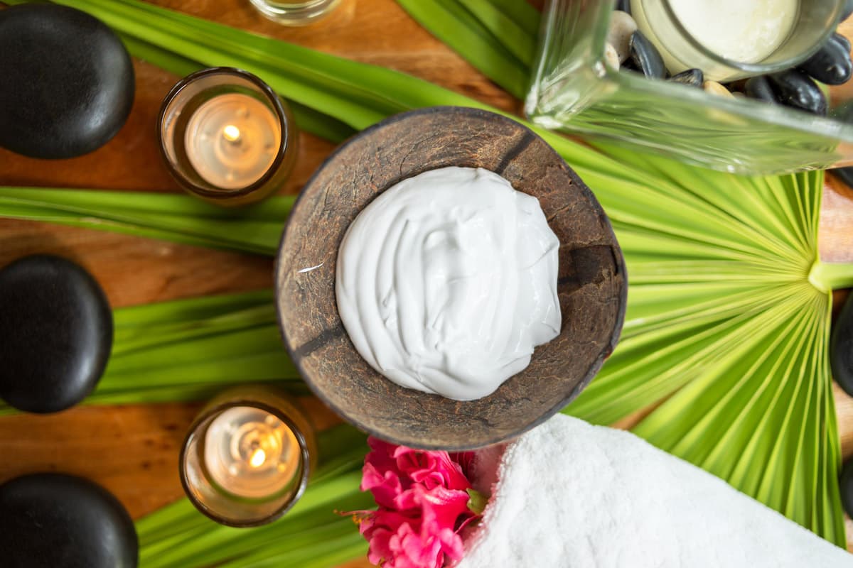 A spa treatment setup featuring a white cream in a coconut shell bowl, surrounded by palm leaves, hot stones, candles, and a white towel.