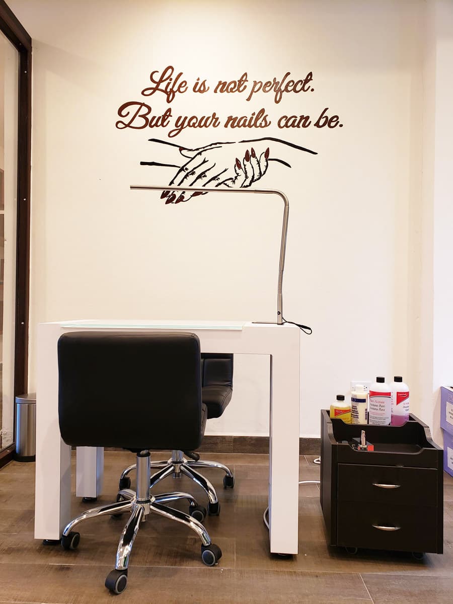 A modern nail salon with a white manicure table and two black swivel chairs. On the wall behind the table is a vinyl decal with the quote "Life is not perfect. But your nails can be." A small, black rolling cart with drawers holds various nail care products. The salon has a clean and minimalist aesthetic.