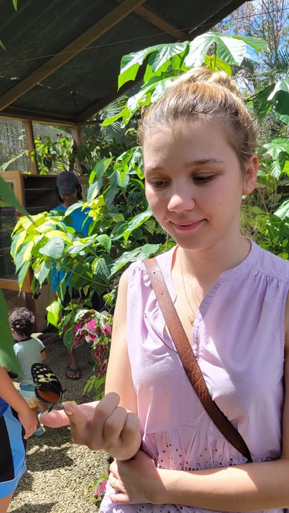 A young lady in a pink top smiles at a butterfly perched on her finger in a butterfly garden.