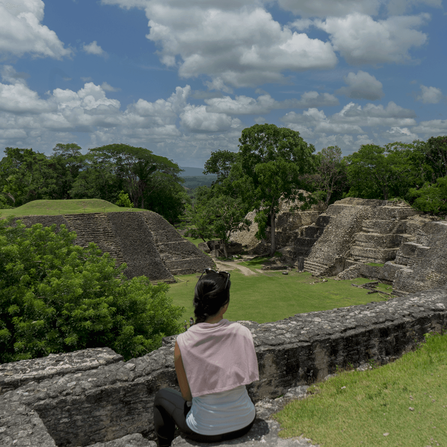 Brunette woman with pink top sitting above the mayan ruins looking down, with green trees and grass and blue sky.