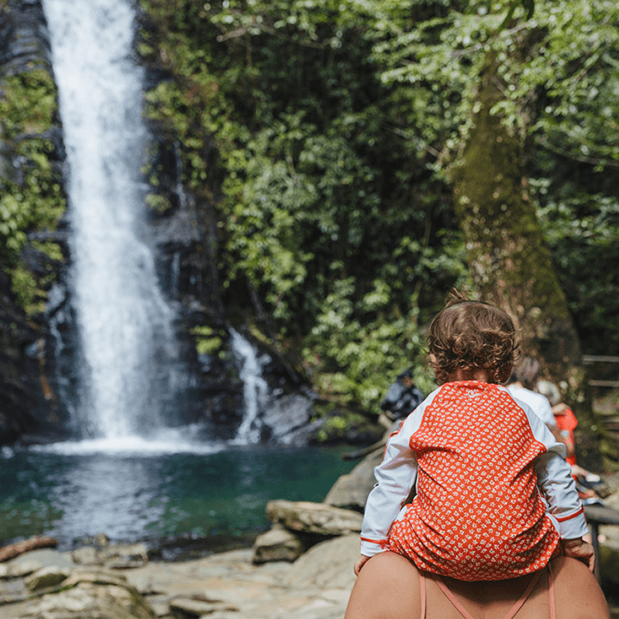 A woman carrying a toddler on her shoulders looks at a waterfall cascading down into a pool surrounded by rocky outcrops.