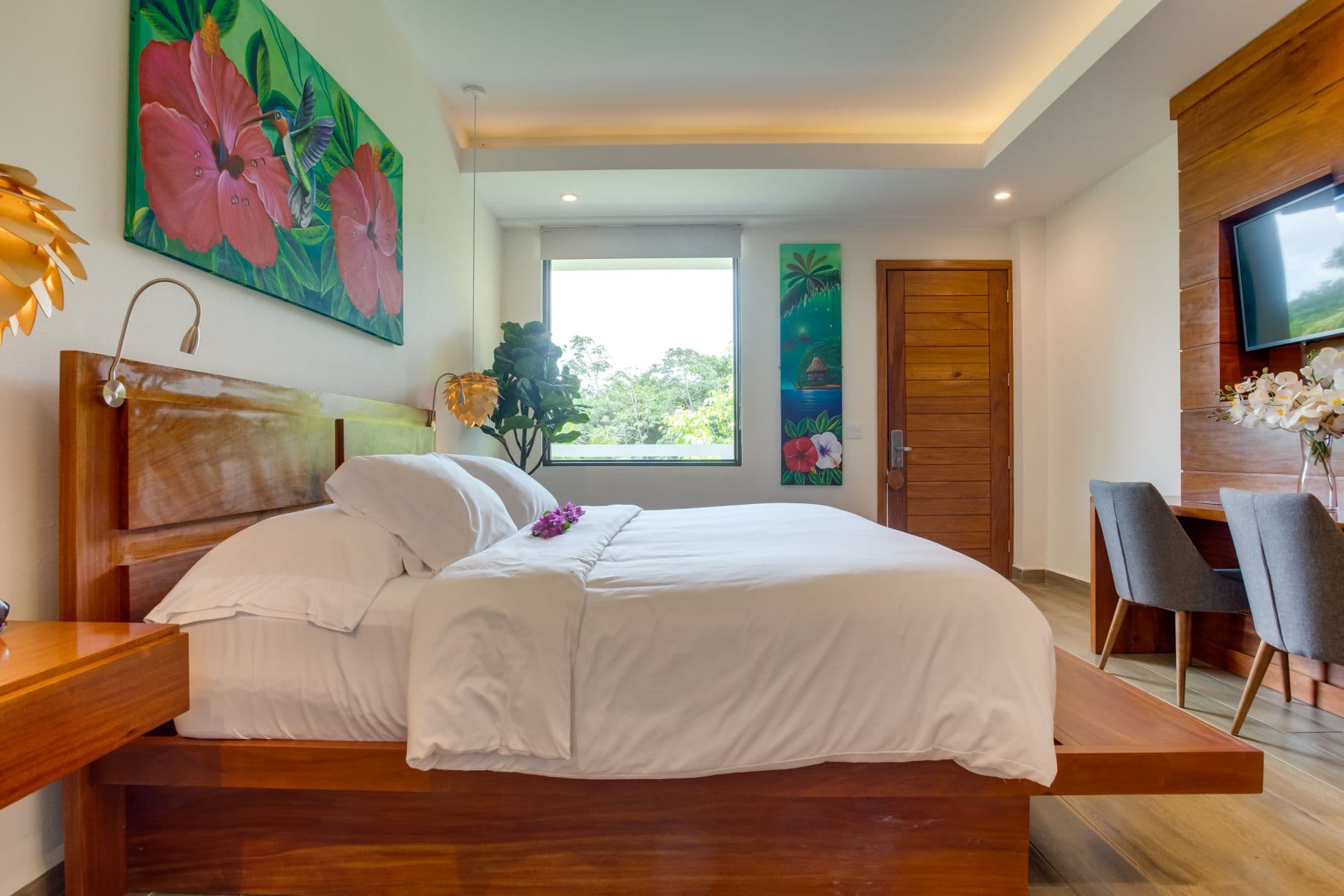 A spacious bedroom with a large bed covered in white sheets. A colorful painting of hummingbirds hangs above the bed, and a large window overlooks a tropical view. A wooden desk with chairs is visible in the corner, and there is a television mounted on the wall. The room has a modern and minimalist design with white walls and wooden accents.