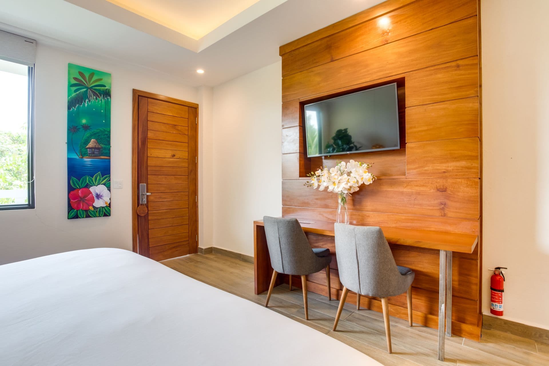 The image shows a hotel room with a modern design. The room features a large bed with white linens, a wooden accent wall with a built-in TV, a wooden desk with two chairs, a colorful painting on the wall, and a large window with natural light.