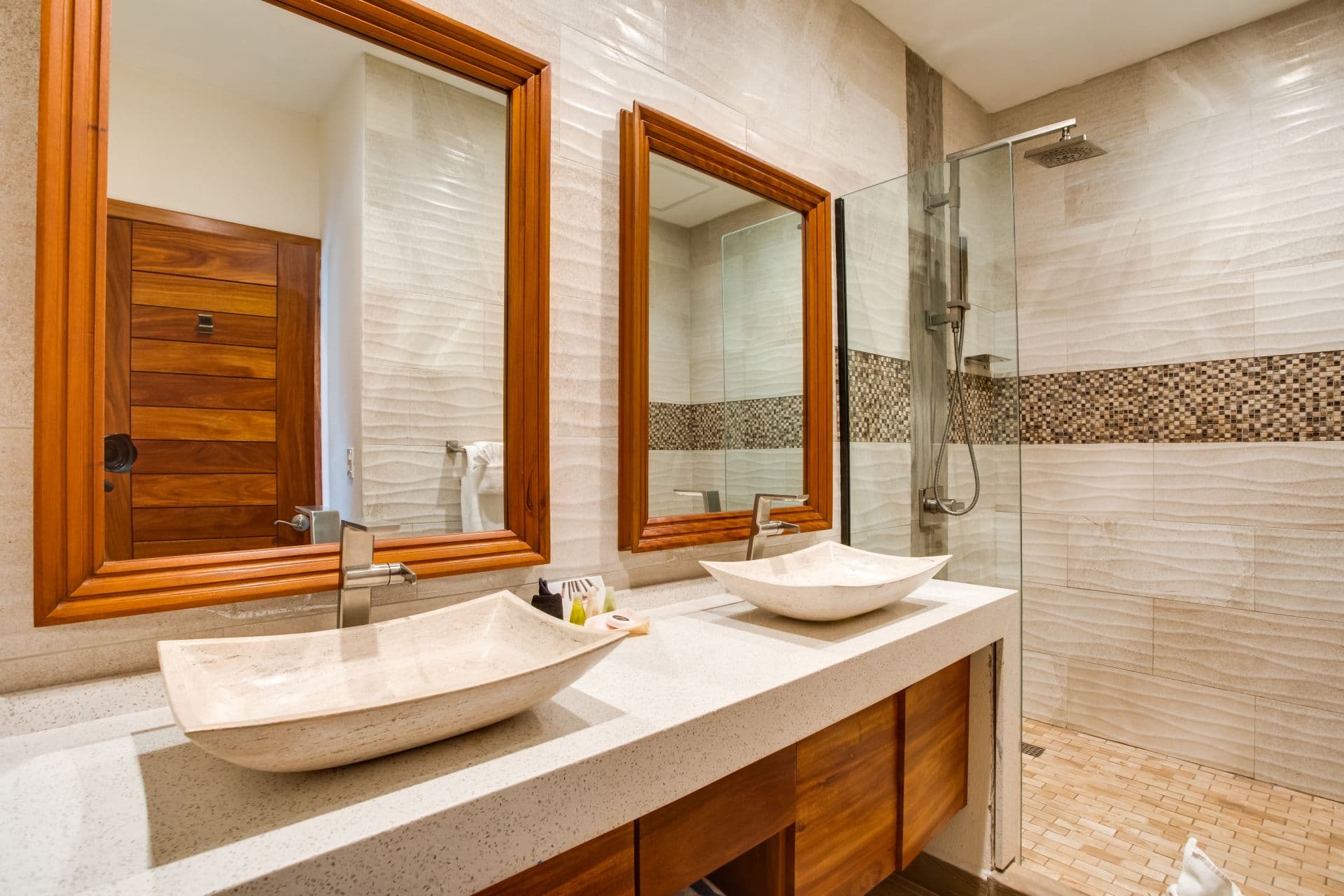 The image shows a modern bathroom with a clean and minimalist design. The bathroom features two rectangular mirrors with wooden frames, two white marble sinks with a wooden countertop, a glass-enclosed shower with a mosaic tile pattern, and a white tiled floor.