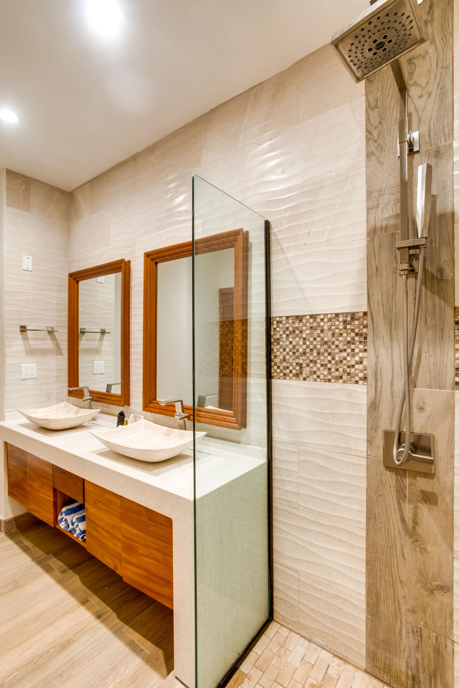 The image shows another bathroom with a similar modern design. This bathroom also features two rectangular mirrors with wooden frames, two white marble sinks with a wooden countertop, a glass-enclosed shower with a mosaic tile pattern, and a white tiled floor. However, this bathroom has a different showerhead and a slightly different layout.