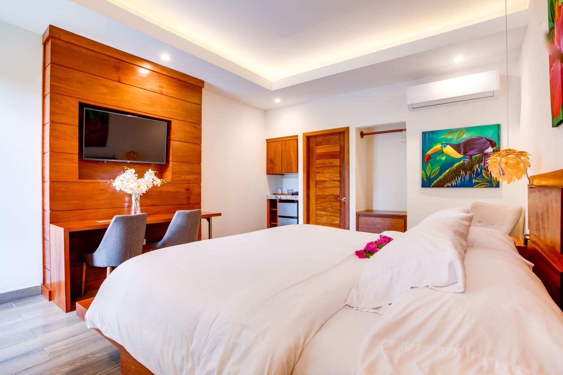 The image shows a modern hotel room with a king-sized bed, a wooden accent wall with a built-in TV, a desk with two chairs, and a colorful painting of a toucan. The room also features a small kitchenette with a mini-fridge and a microwave.