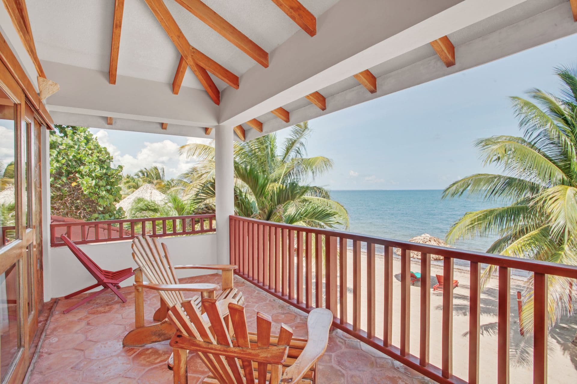 A spacious balcony with two wooden Adirondack chairs overlooking a sandy beach and the ocean. The balcony has a tiled floor and a wooden railing. There are palm trees visible on the beach, and the sky is blue and sunny.