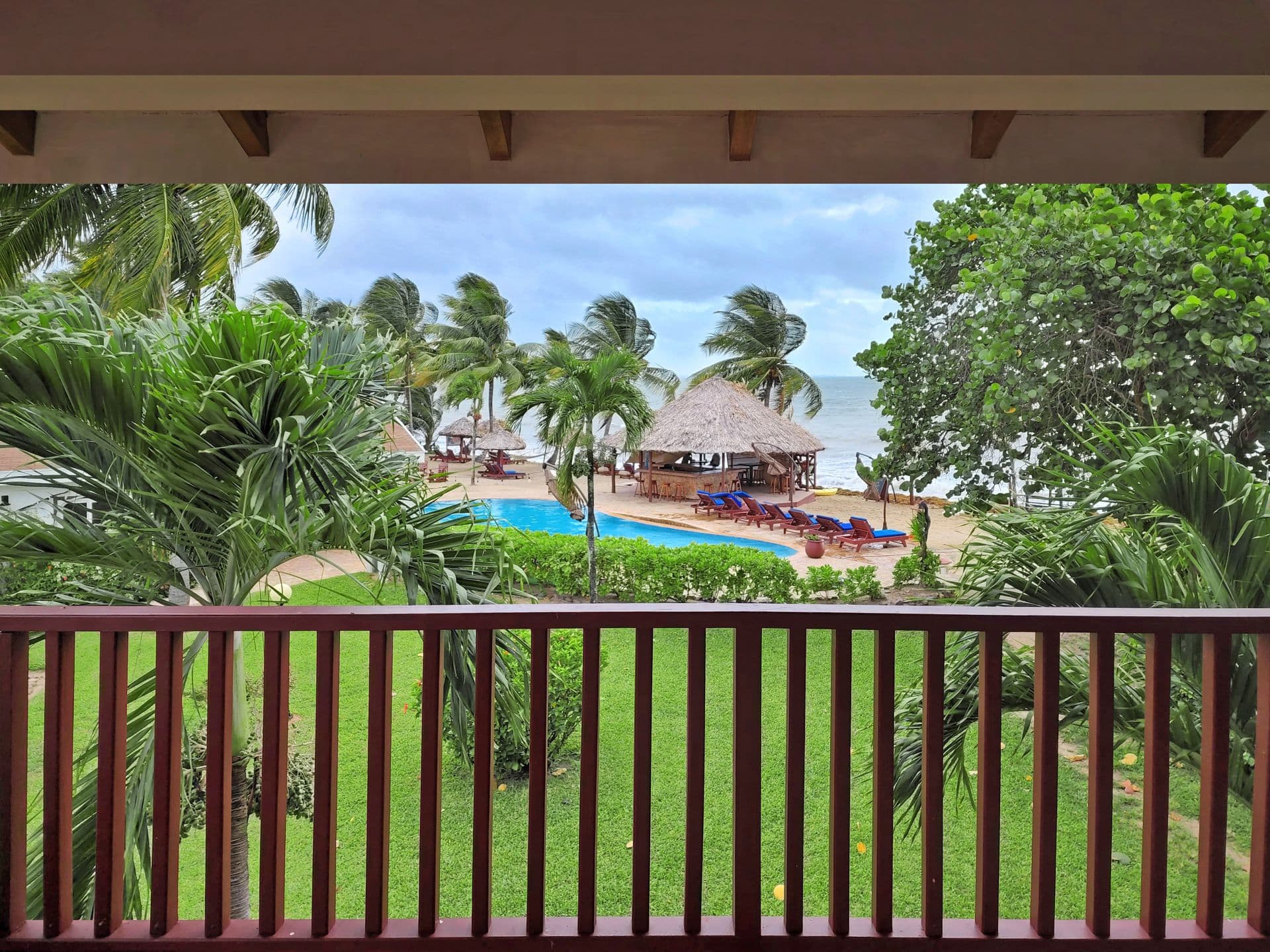A balcony overlooking a tropical resort with a swimming pool, bar, and palm trees. The ocean is visible in the background, and the sky is cloudy.
