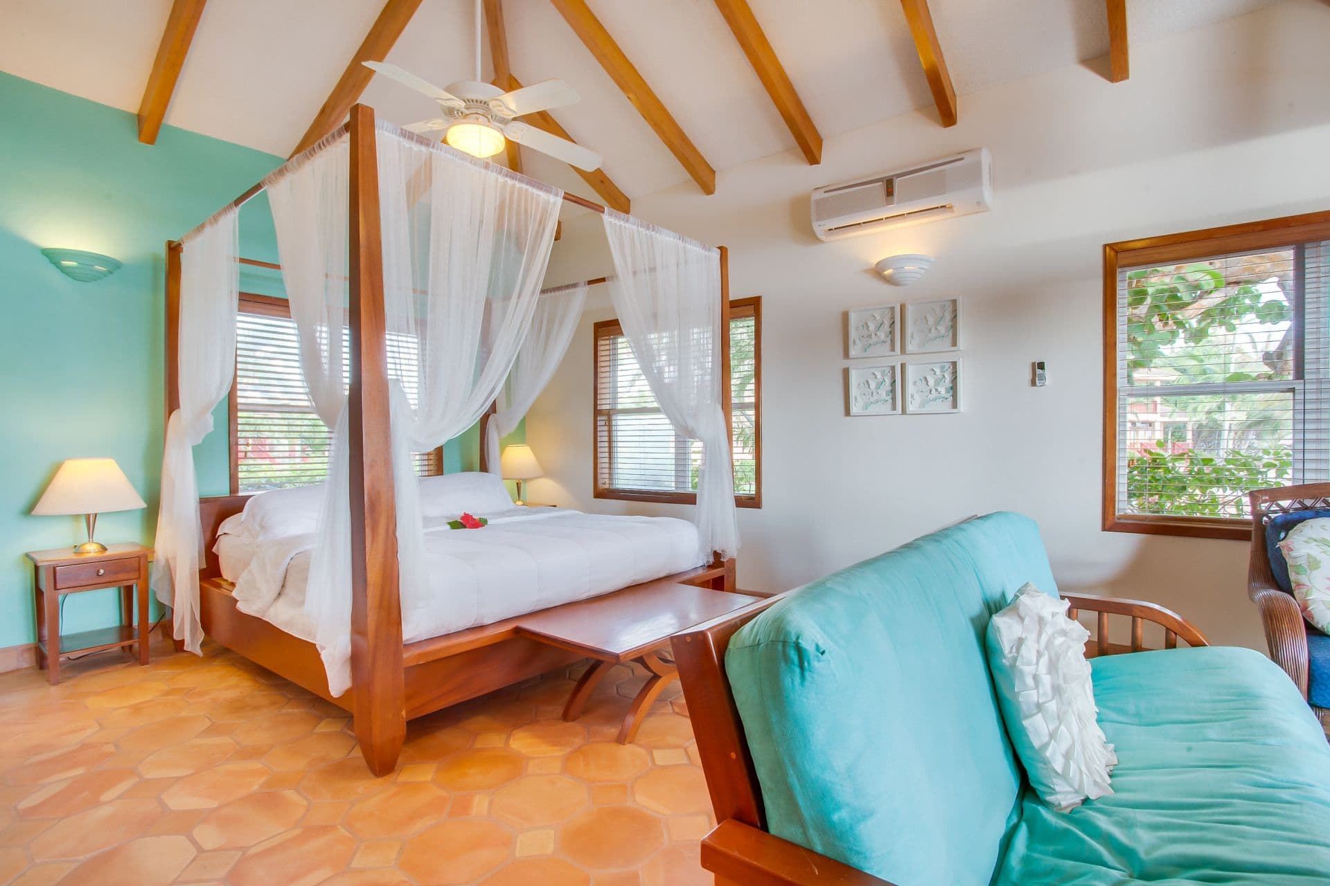 A spacious bedroom with a four-poster bed covered in white sheets. The bed has sheer white curtains hanging from the canopy. A ceiling fan hangs from the center of the room, and there is an air conditioning unit in the background. Nightstands with lamps flank the bed, and a wooden bench sits at the foot. A small sofa sits in the corner of the room, and a coffee table is placed between the sofa and the bed. The room has a tiled floor and a wooden closet door. The walls are painted a light blue color.