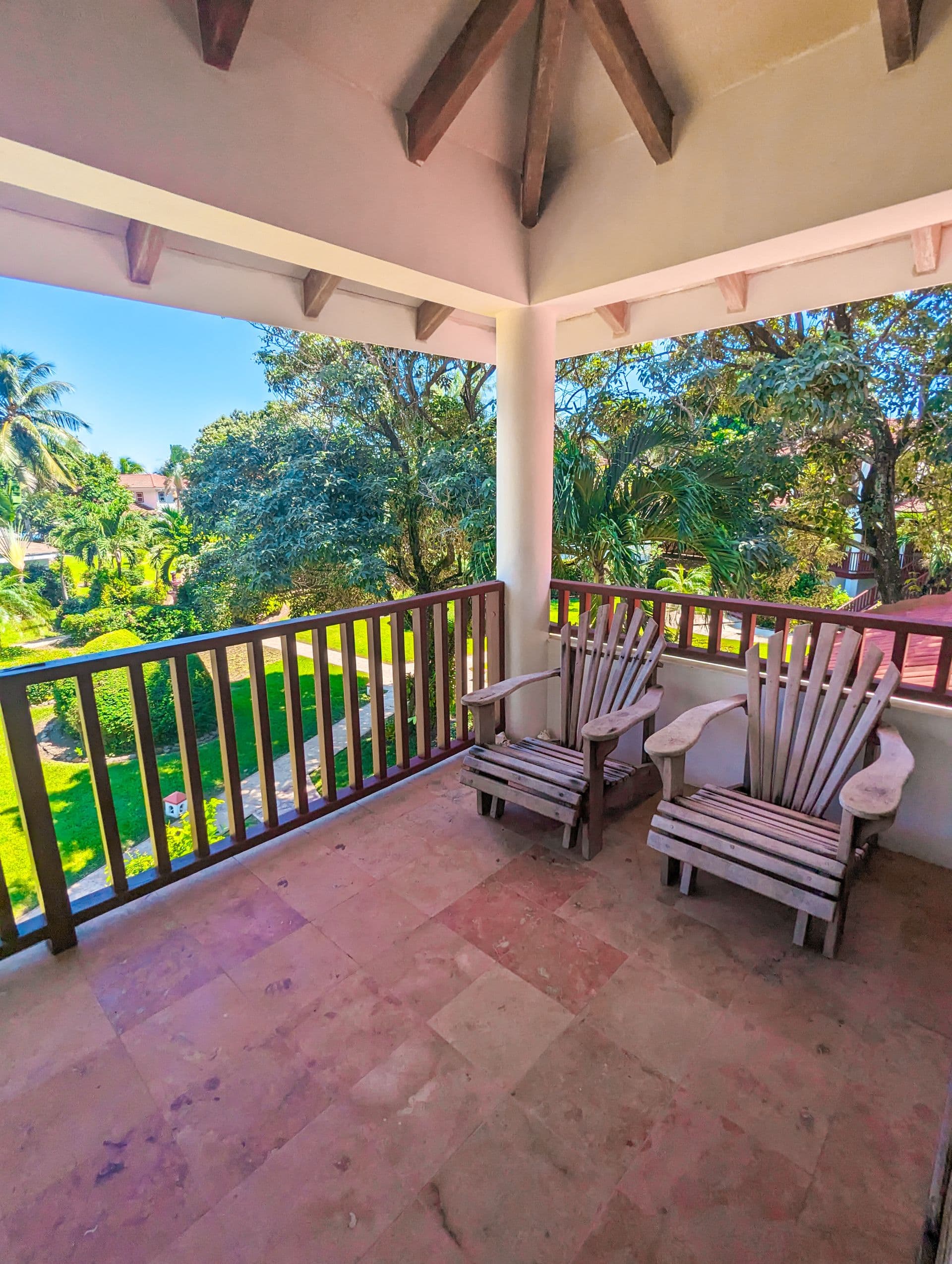 A spacious balcony with two wooden Adirondack chairs overlooking a lush tropical garden. The balcony has a tiled floor and a wooden railing. There are palm trees and other tropical plants visible in the background. The sky is blue and sunny